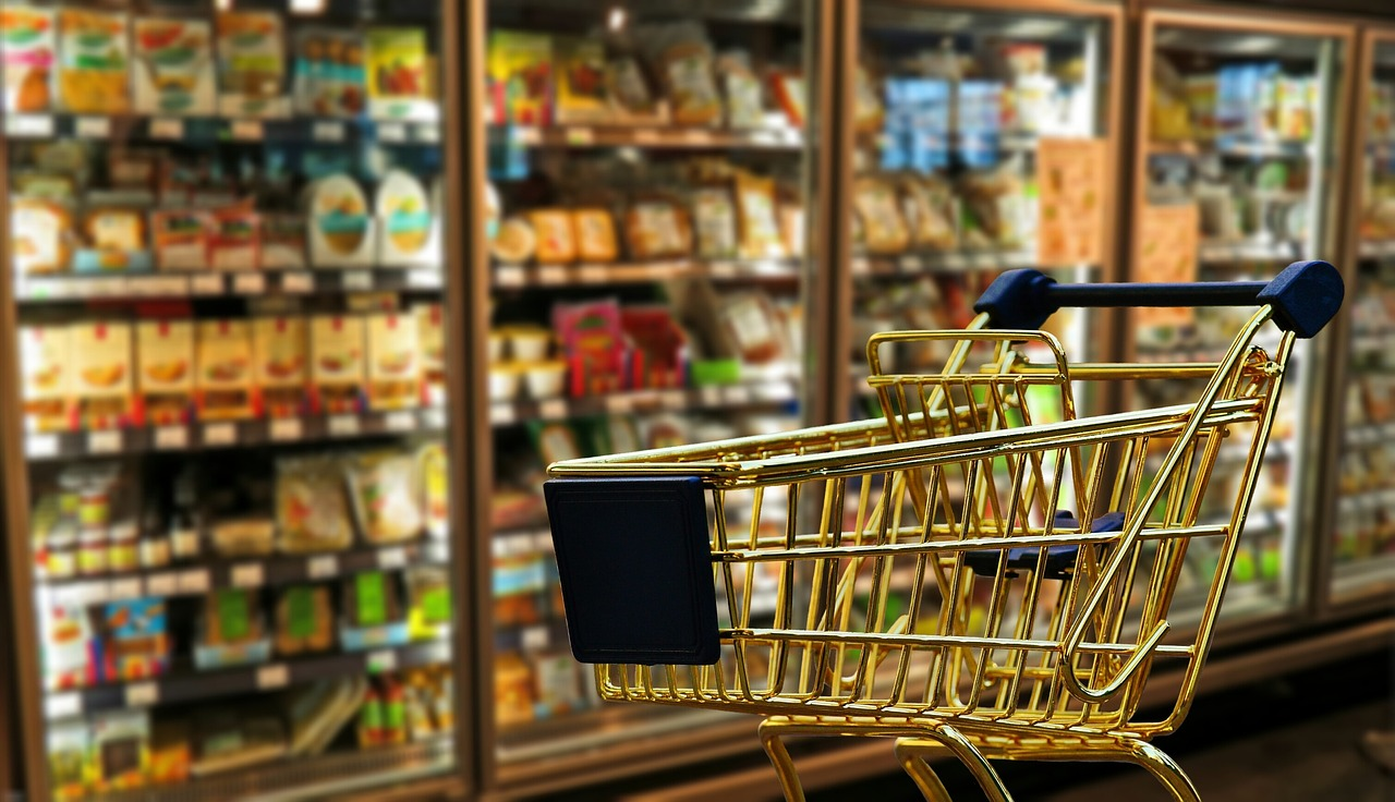 Shopping cart in the foreground with various grocery items behind.
