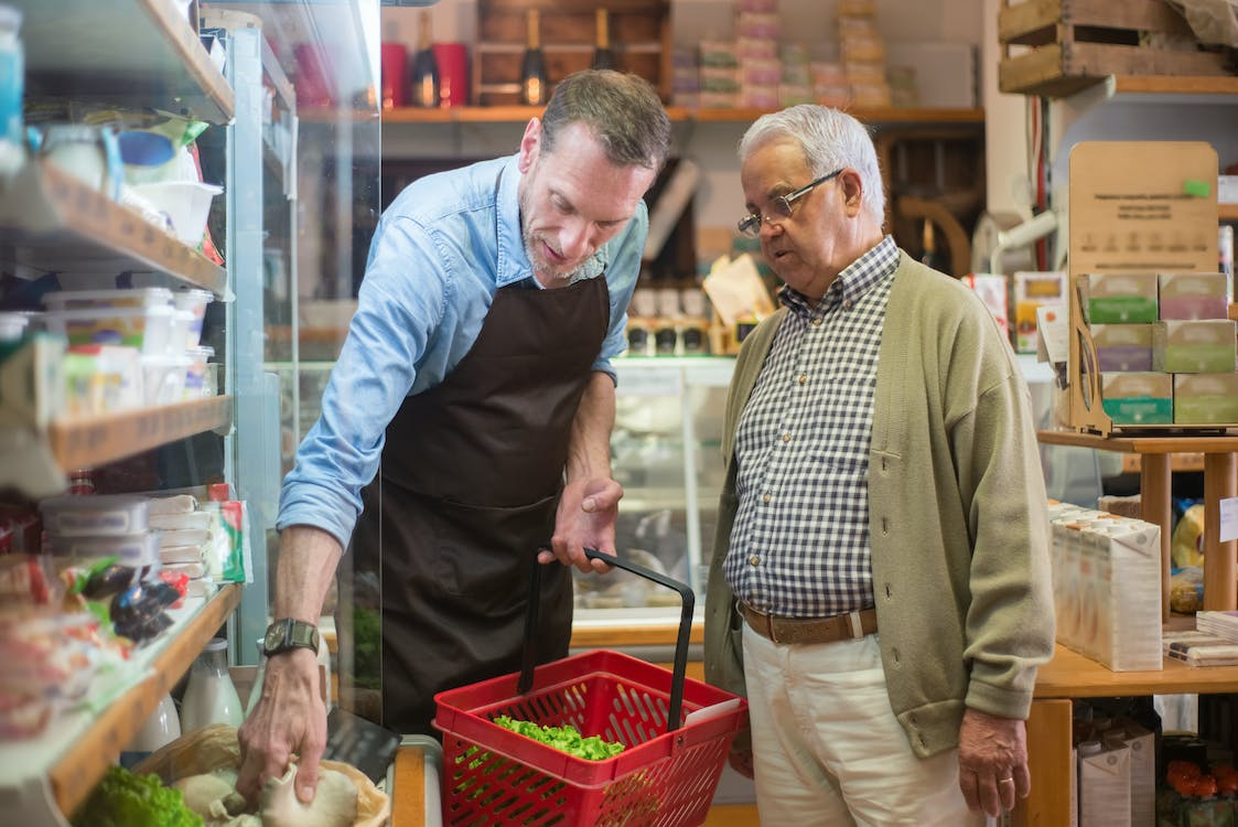 grocer assisting a senior customer with produce selection in a health food store.